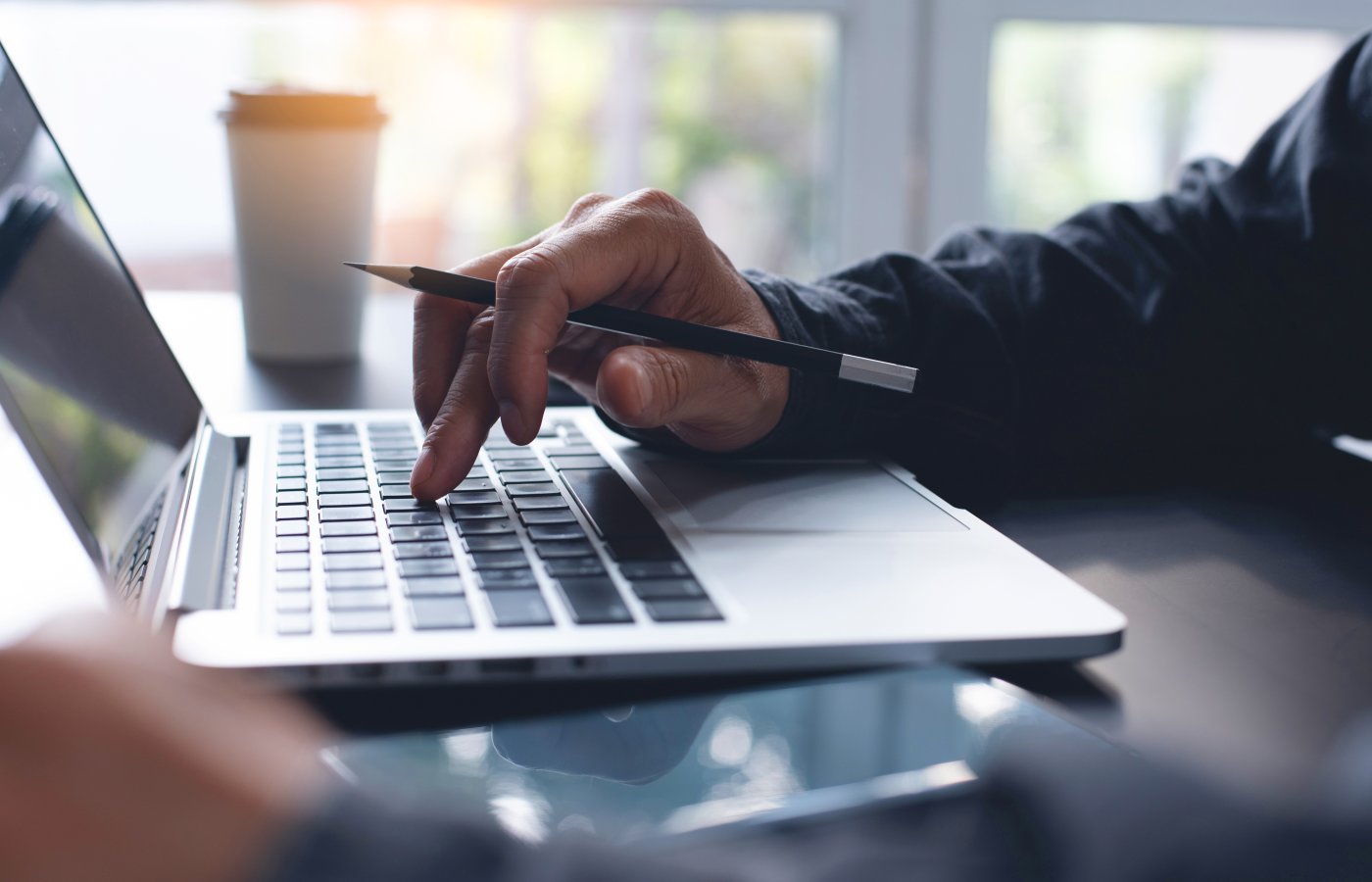 close up of hands typing on a laptop keyboard with a pencil and warm beverage cup. 