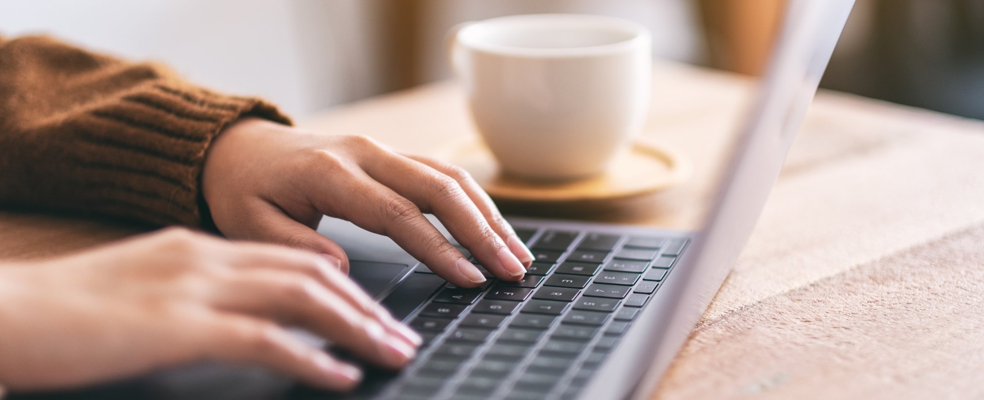 close up of hands typing on laptop with a coffee cup next to it. 